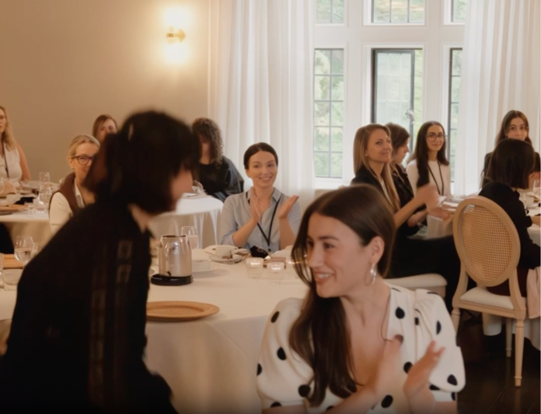 Attendees seated at tables during an indoor studio event, applauding while listening to a speaker.