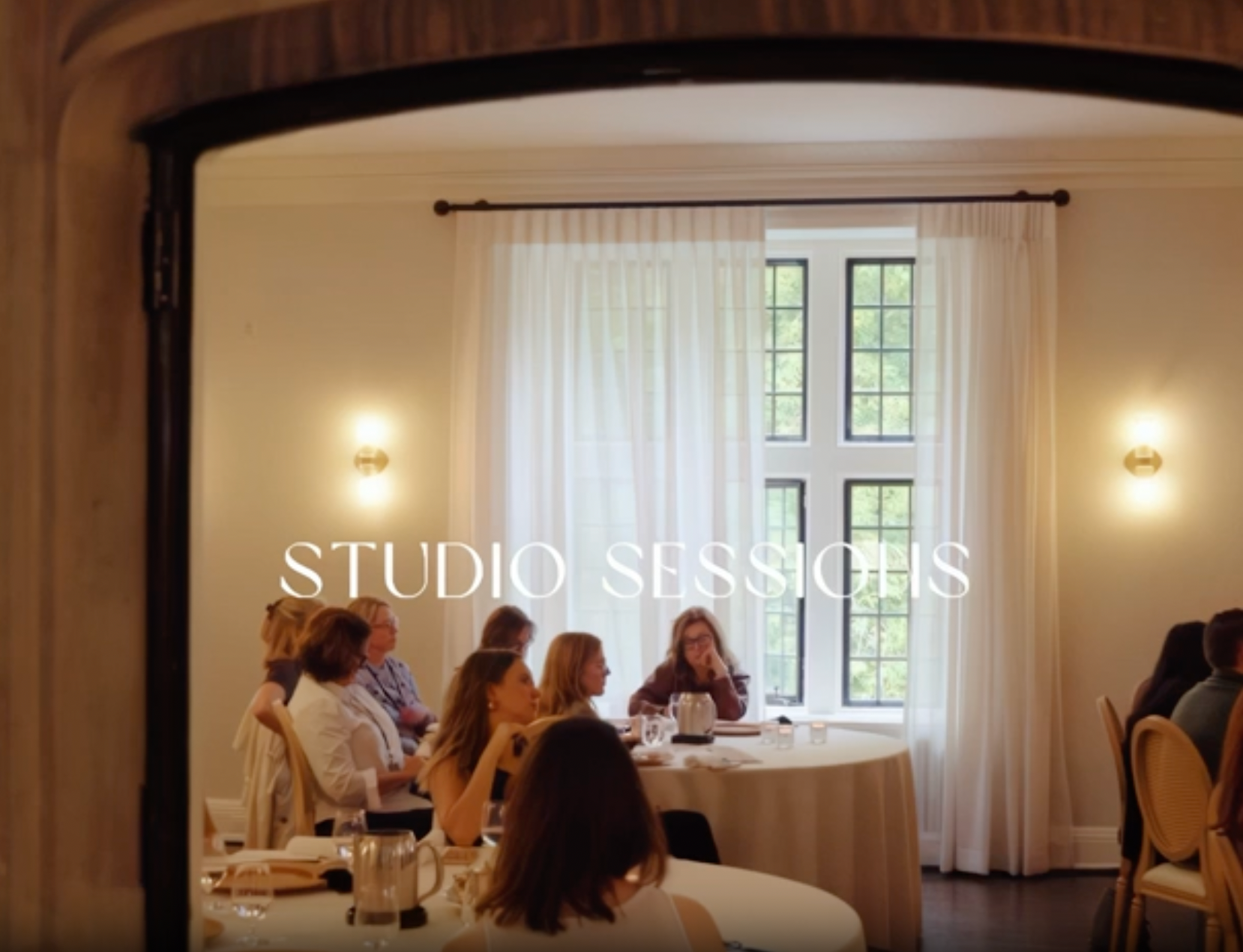 Attendees seated at round tables during an indoor studio session event, listening and taking notes in a bright meeting space.